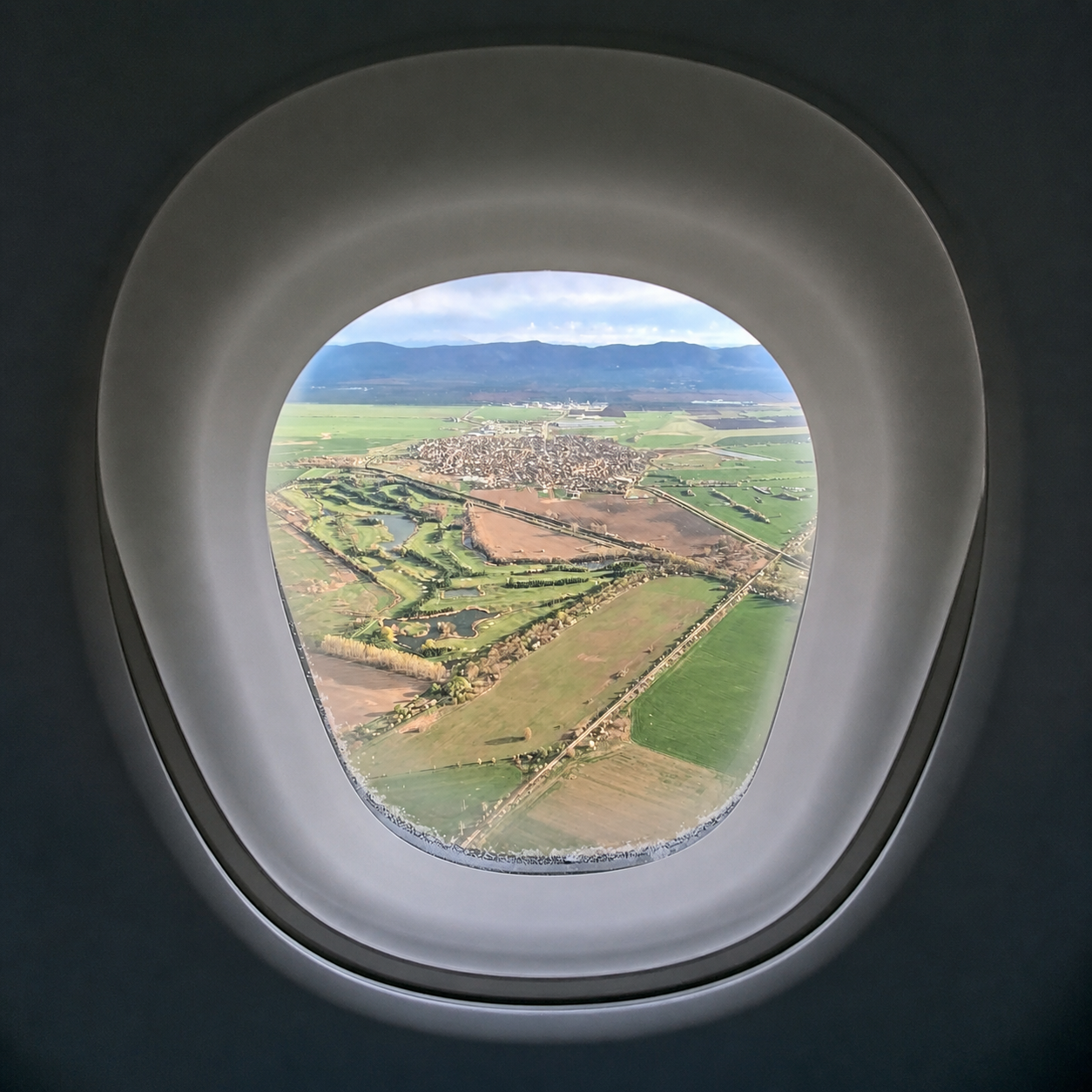 Aerial view through an airplane window of St. Sofia Golf Club near Sofia, Bulgaria, showing green fairways, water hazards, tree-lined golf paths, surrounding farmland, and distant mountain ranges under soft daylight.